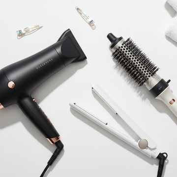 A flat-lay arrangement of hair styling tools on a white background, including a black hair dryer with rose gold accents, a white round brush attachment, a white flat iron, and a white towel with small hair clips.